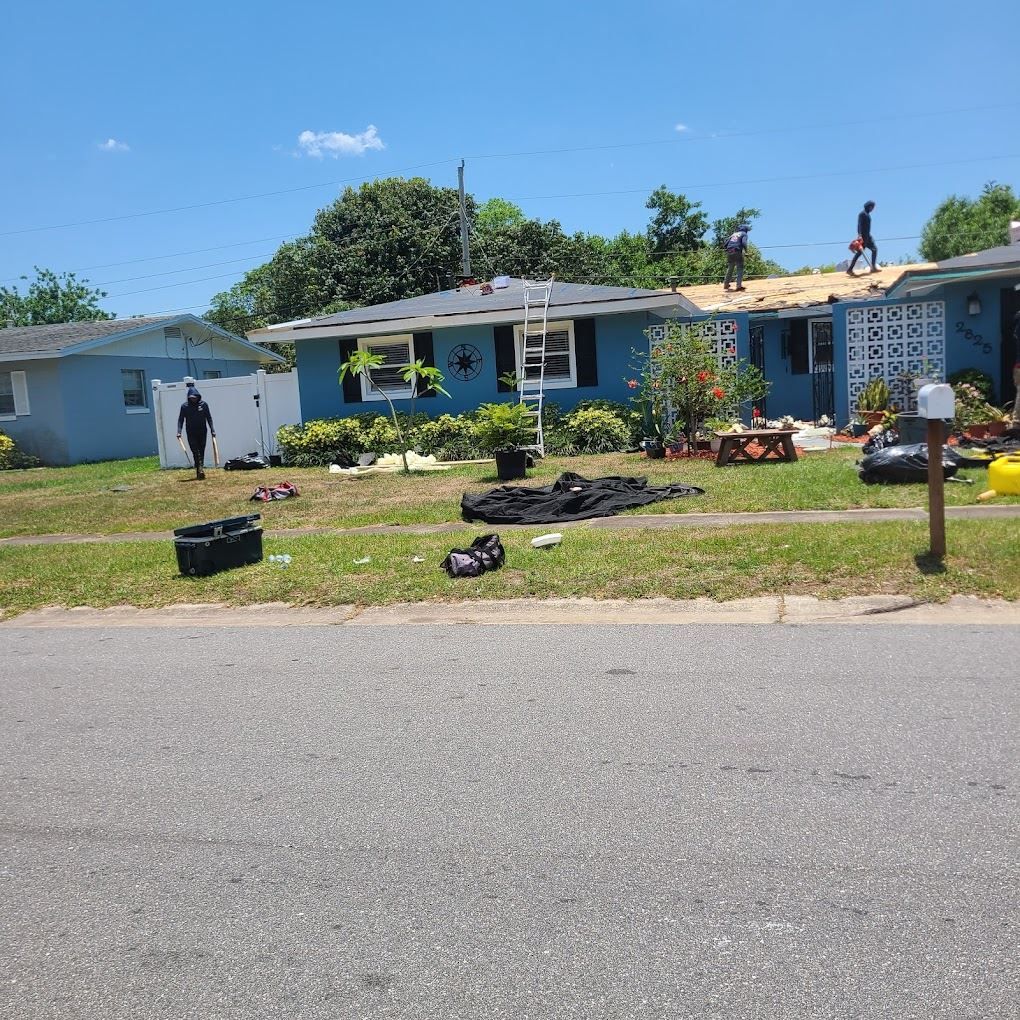 Workers repair a roof on a blue house in a residential neighborhood under a sunny sky.