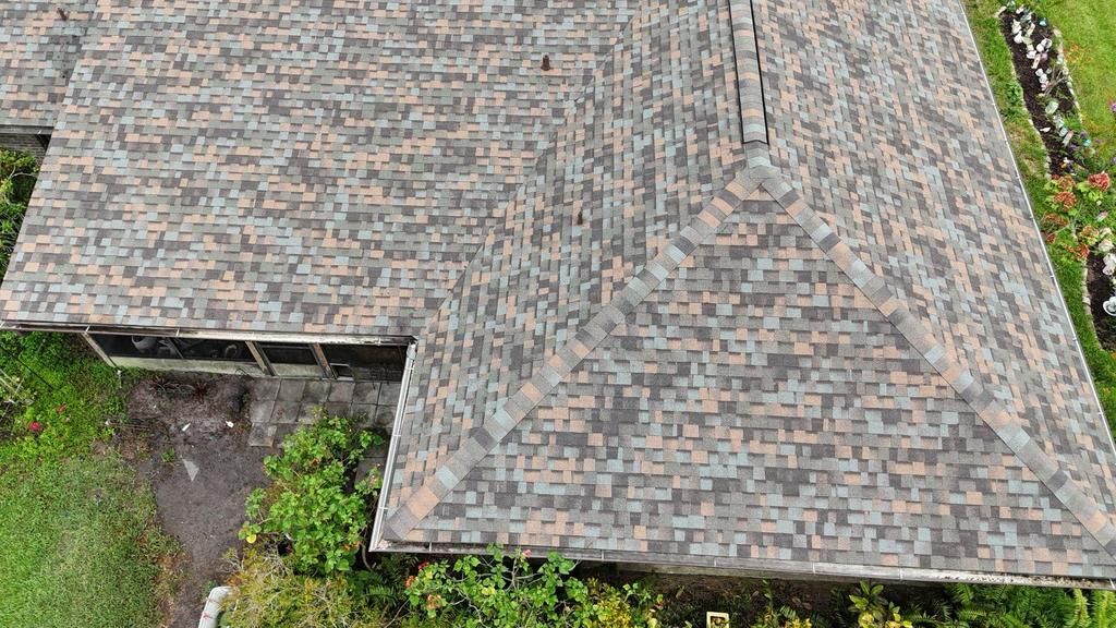 Aerial view of an L-shaped residential roof with variegated brown and grey shingles surrounded by greenery.