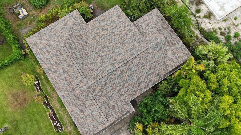 An aerial view of a house roof with textured, multi-toned brown shingles surrounded by green trees and a grassy lawn.