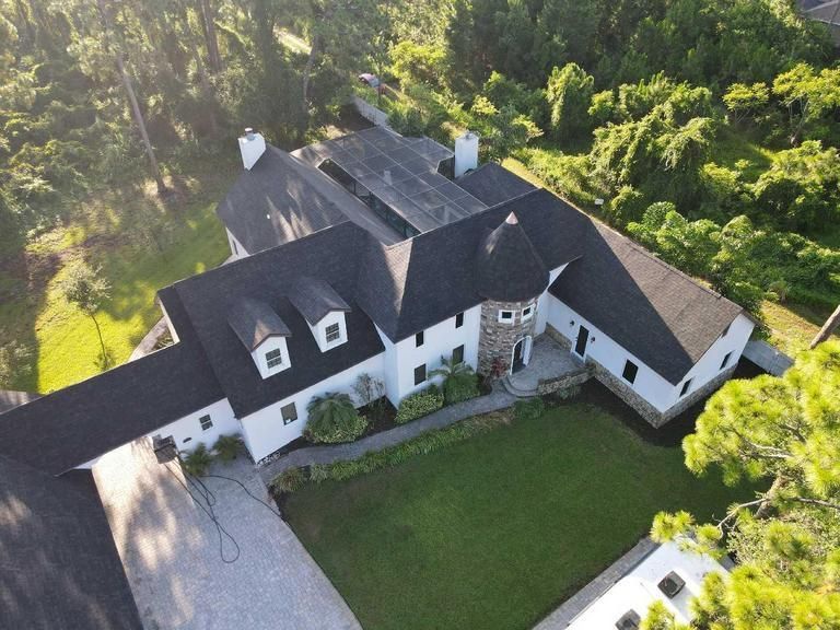 An aerial view of a white, multi-level house with a dark roof and stone turret, surrounded by trees and a green lawn.