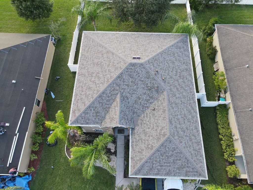 Aerial view of a residential house with a gray shingled roof, surrounded by green lawns and a white fence.