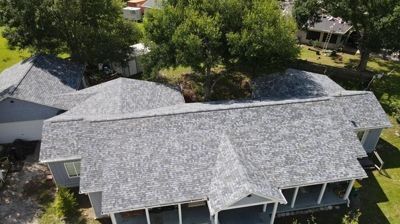 An aerial view of a gray shingled roof on a house with a covered front porch, surrounded by green trees and lawn.