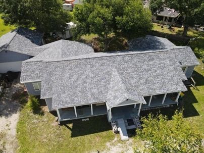 Aerial view of a gray-roofed residential home with a wrap-around porch, surrounded by green trees and lawn.
