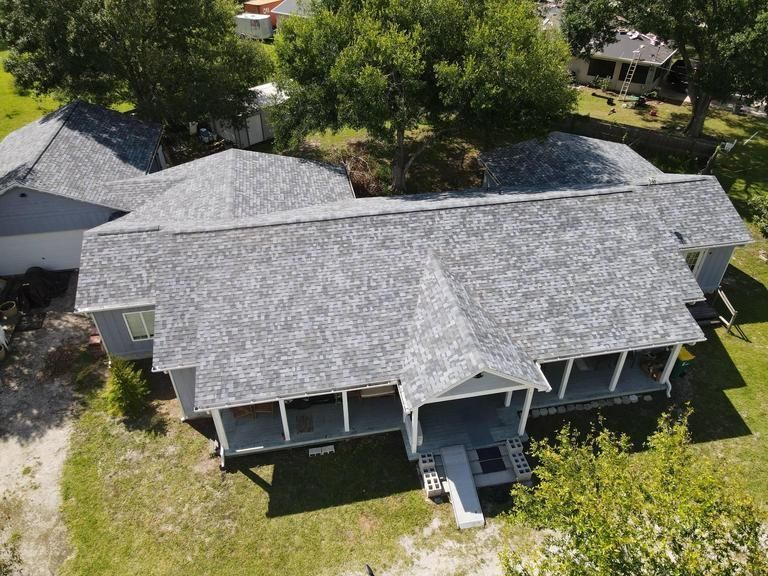 An aerial view of a residential house with a gray shingled roof, a covered front porch, and surrounding green lawn and trees.