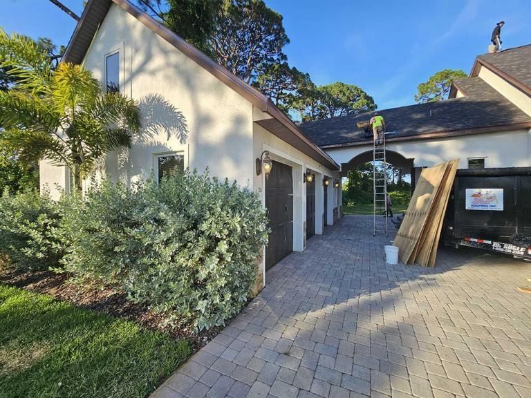 Construction workers on the roof of a house with a paved driveway, a large dumpster, and landscaping in bright sunlight.