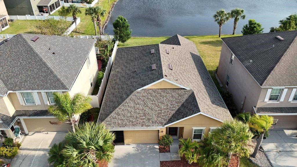 Aerial view of three suburban homes with asphalt shingle roofs, situated near a small lake on a sunny day.