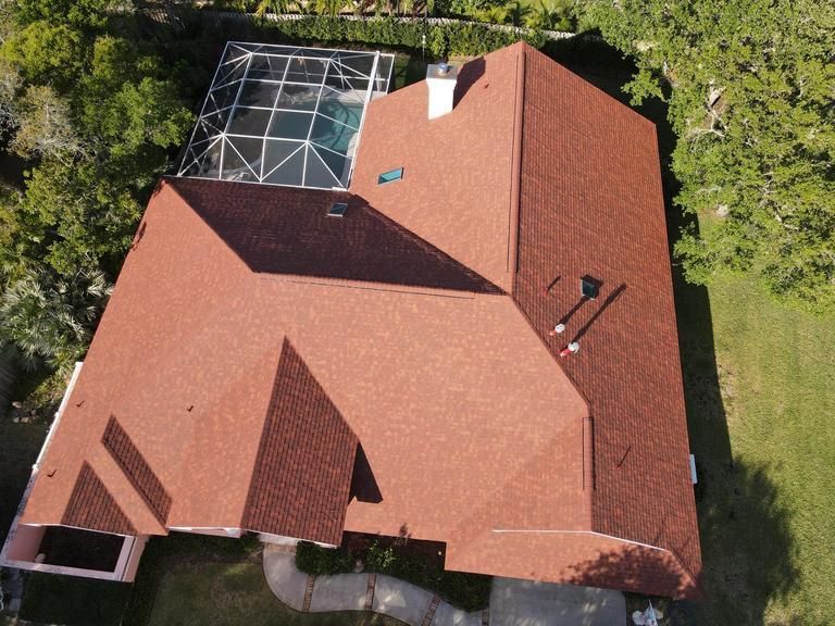 An aerial view of a reddish-brown shingled house roof with a pool enclosure in a wooded, suburban setting.