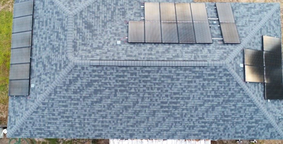 An aerial view of a dark grey shingled residential roof with multiple black solar panel arrays installed on various slopes.