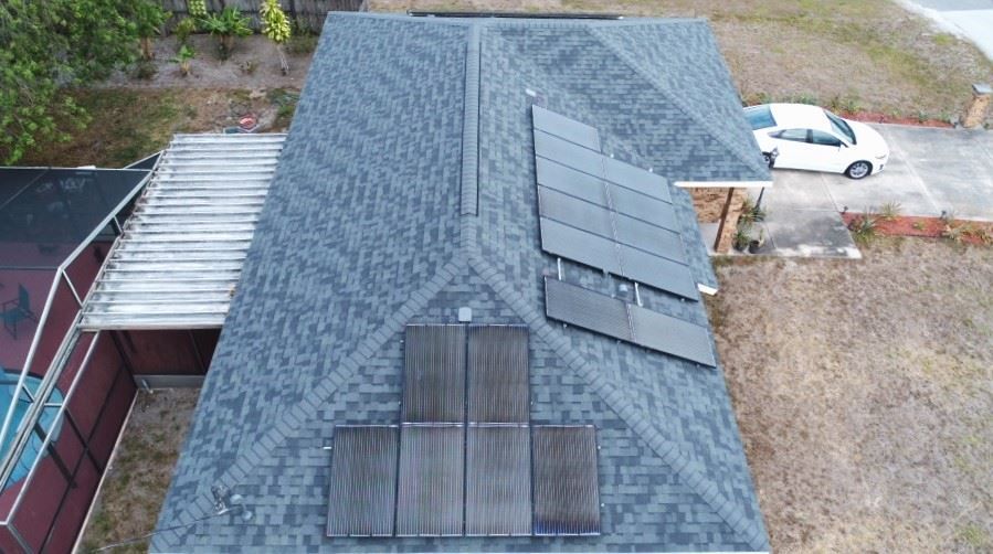 An aerial view of a dark shingled residential roof featuring solar electric panels and dark solar pool heating panels.