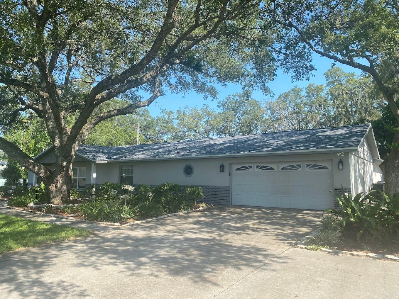Single-story gray house with a two-car garage and large oak trees in a sunny front yard.