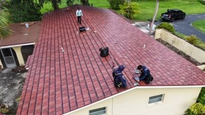 Three roofers work on a newly installed red shingle roof of a house, seen from an elevated angle.