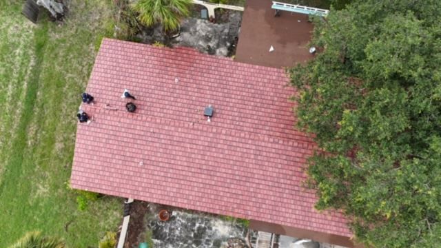 An aerial view of a red roof with two people working on the left side, surrounded by trees and yard areas.