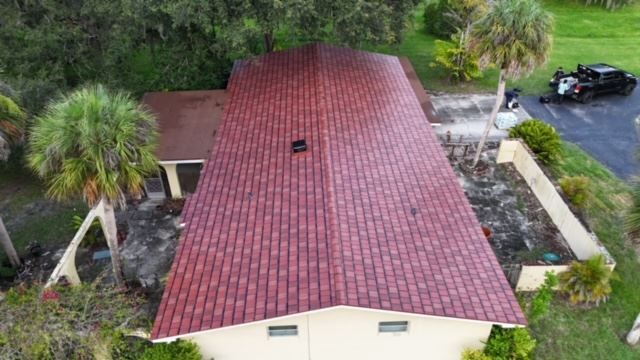 A high-angle view of a rectangular house with a deep red shingled roof, surrounded by palm trees and a paved driveway.