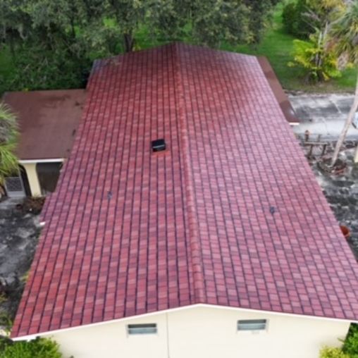 An aerial view of a rectangular house with a shingled roof in shades of red and brown, surrounded by trees and greenery.