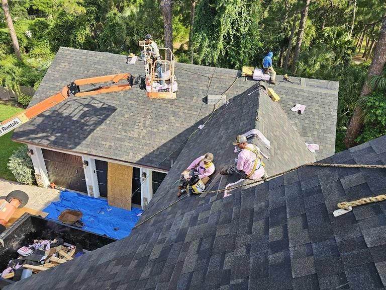 Workers using a lift and safety gear to install new asphalt shingles on a multi-sloped residential roof.