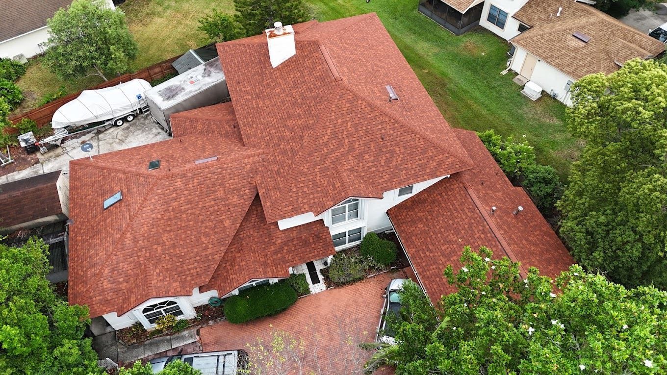 Aerial view of a white home with a reddish-brown shingled roof, featuring a brick driveway and surrounding green trees.