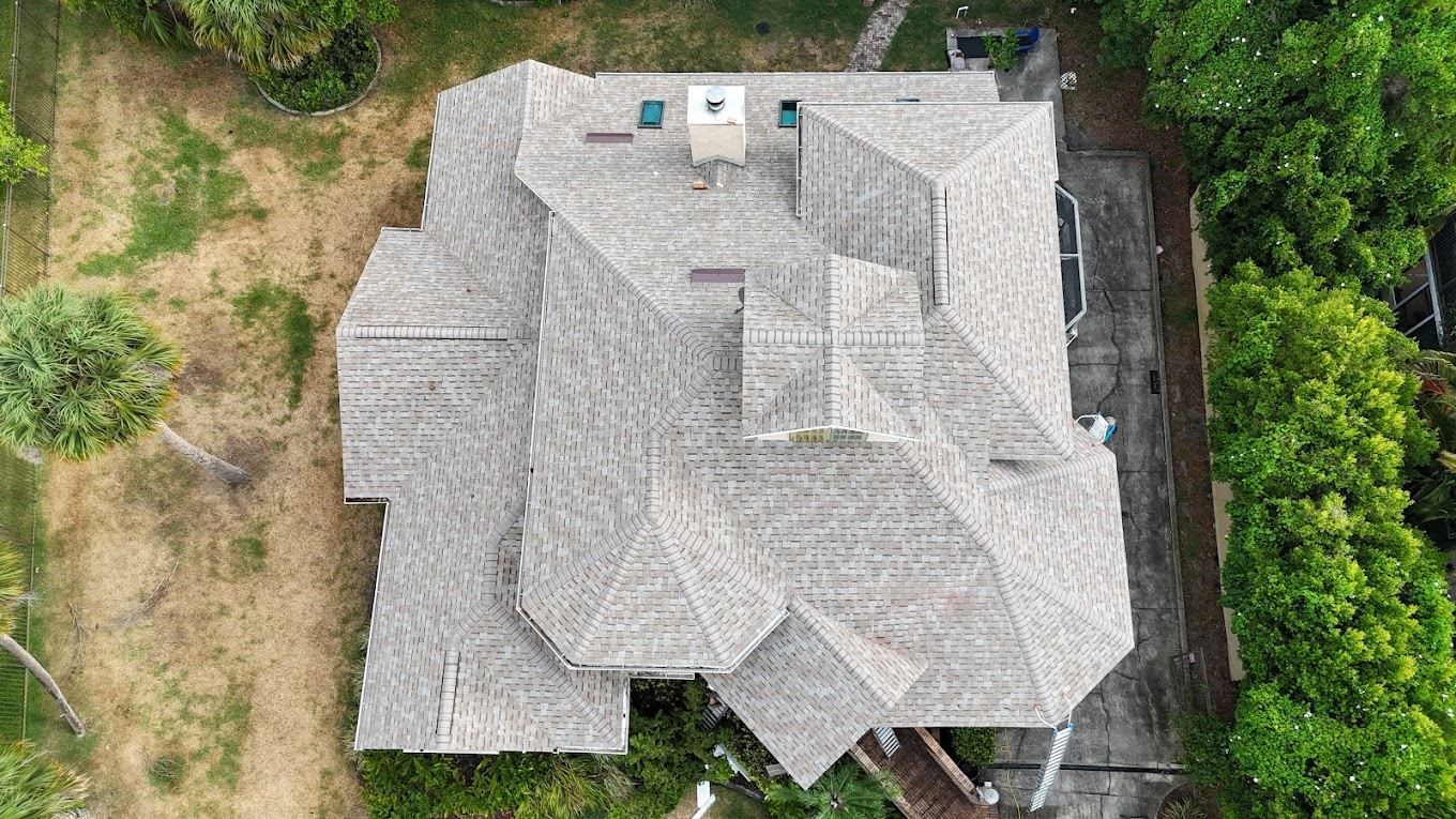 A top-down aerial view of a house with a complex, shingled roof, surrounded by trees and a paved driveway.