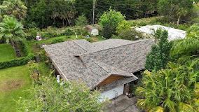 An aerial view of a residential house with a gray shingled roof, surrounded by lush green trees and a small yard.