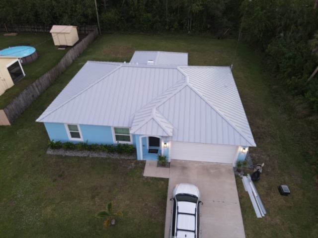 A blue, single-story house with a light gray metal roof, viewed from an elevated angle, with a car in the driveway.