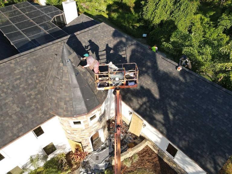 An aerial view of a worker in a cherry picker lift repairing the roof of a stone and white home surrounded by trees.