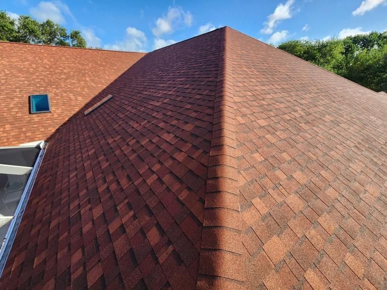 A high-angle view of a red-toned asphalt shingle roof with a prominent ridge line and a small skylight against a blue sky.