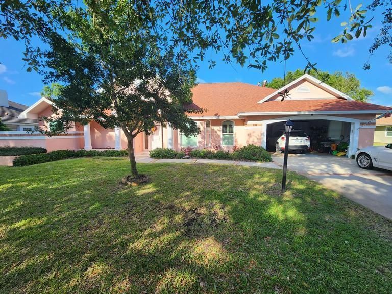 A one-story peach-colored house with a red roof, a tree in the front lawn, and a garage containing a parked car.