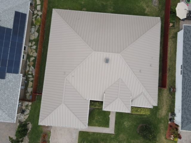 An aerial view of a light-colored, multi-pitched metal roof on a residential home surrounded by lawns and neighboring houses.