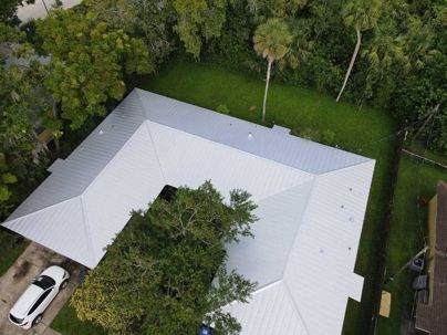 Aerial view of a U-shaped house with a light gray metal roof, surrounded by trees and a green lawn.