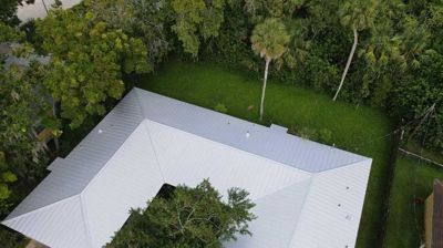 Aerial view of a house with a gray metal roof, surrounded by a lush green lawn and trees.
