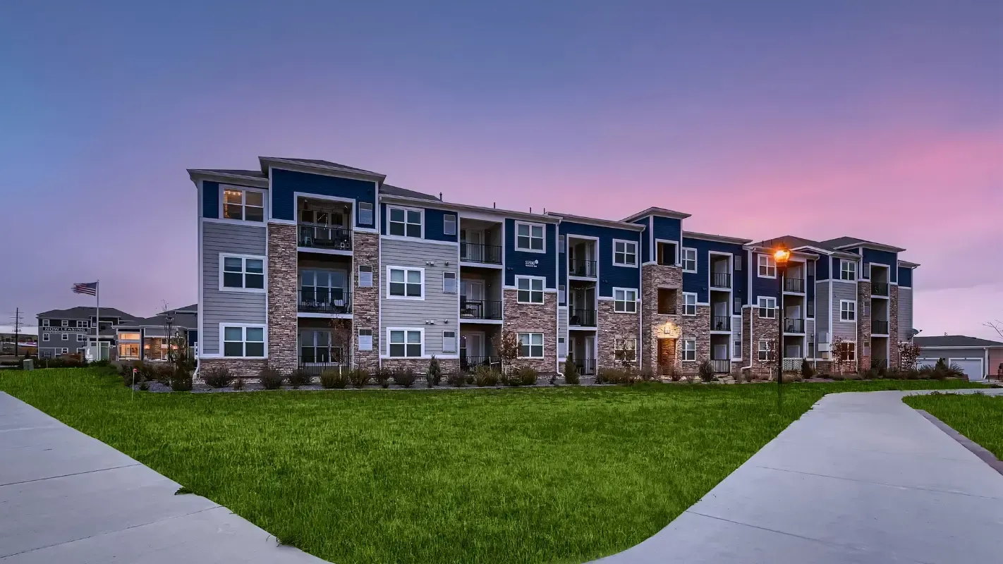 Exterior view of a multi-unit apartment building with blue siding, stone accents, and a lawn at dusk.