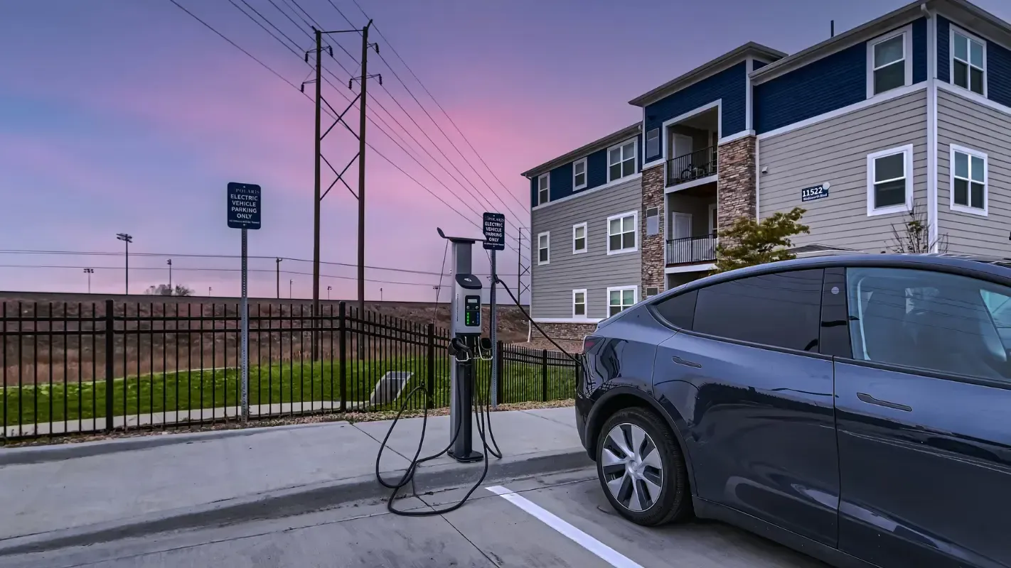 Electric vehicle charging station beside a parked car in front of a modern apartment building at sunset.