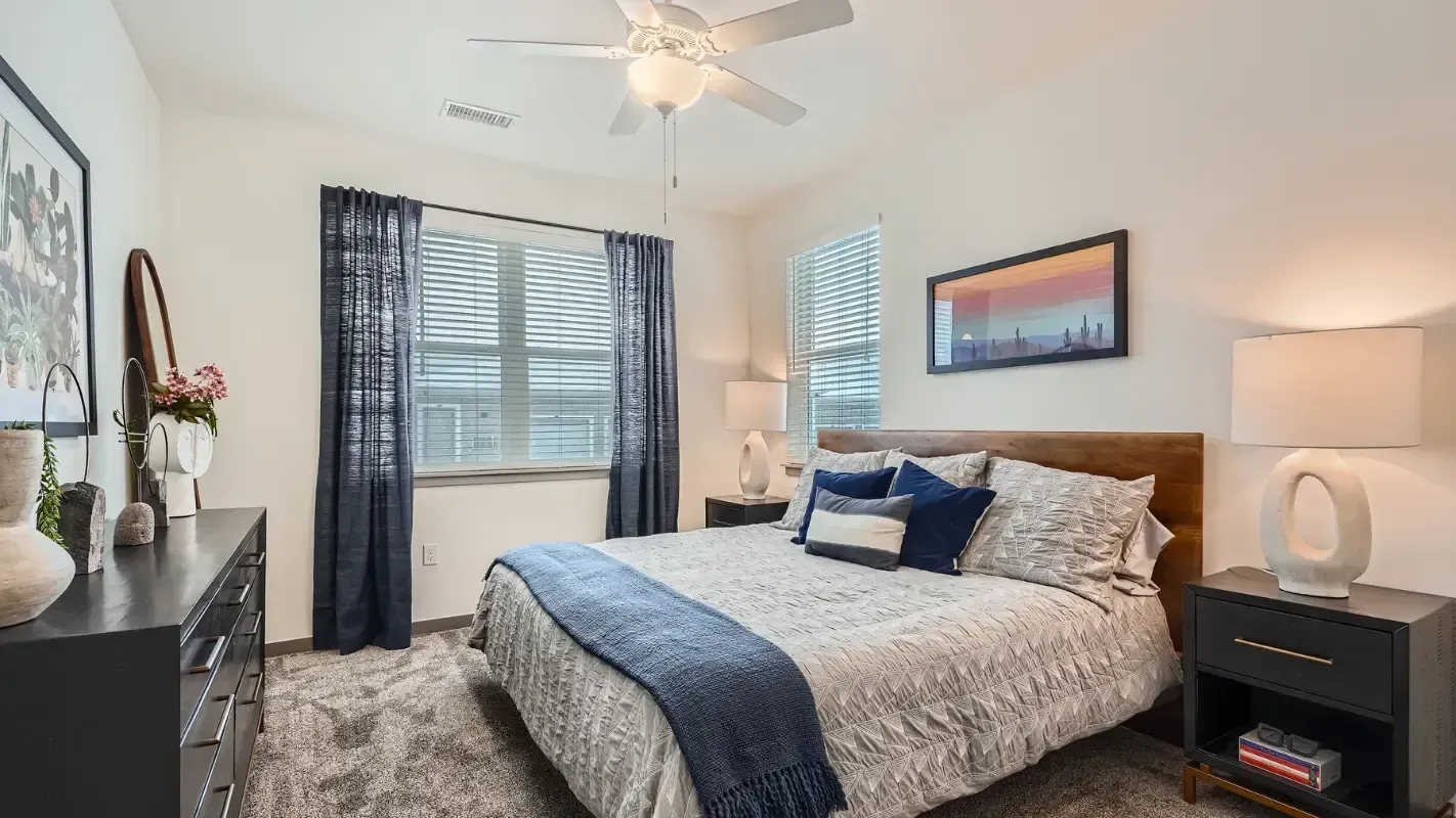 Bedroom in a modern apartment with a bed, nightstands, dresser, and a window with dark curtains.