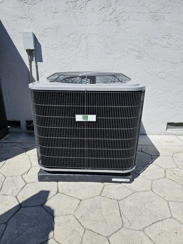 An outdoor air conditioning unit against a light gray wall on a patterned stone patio, casting a shadow.