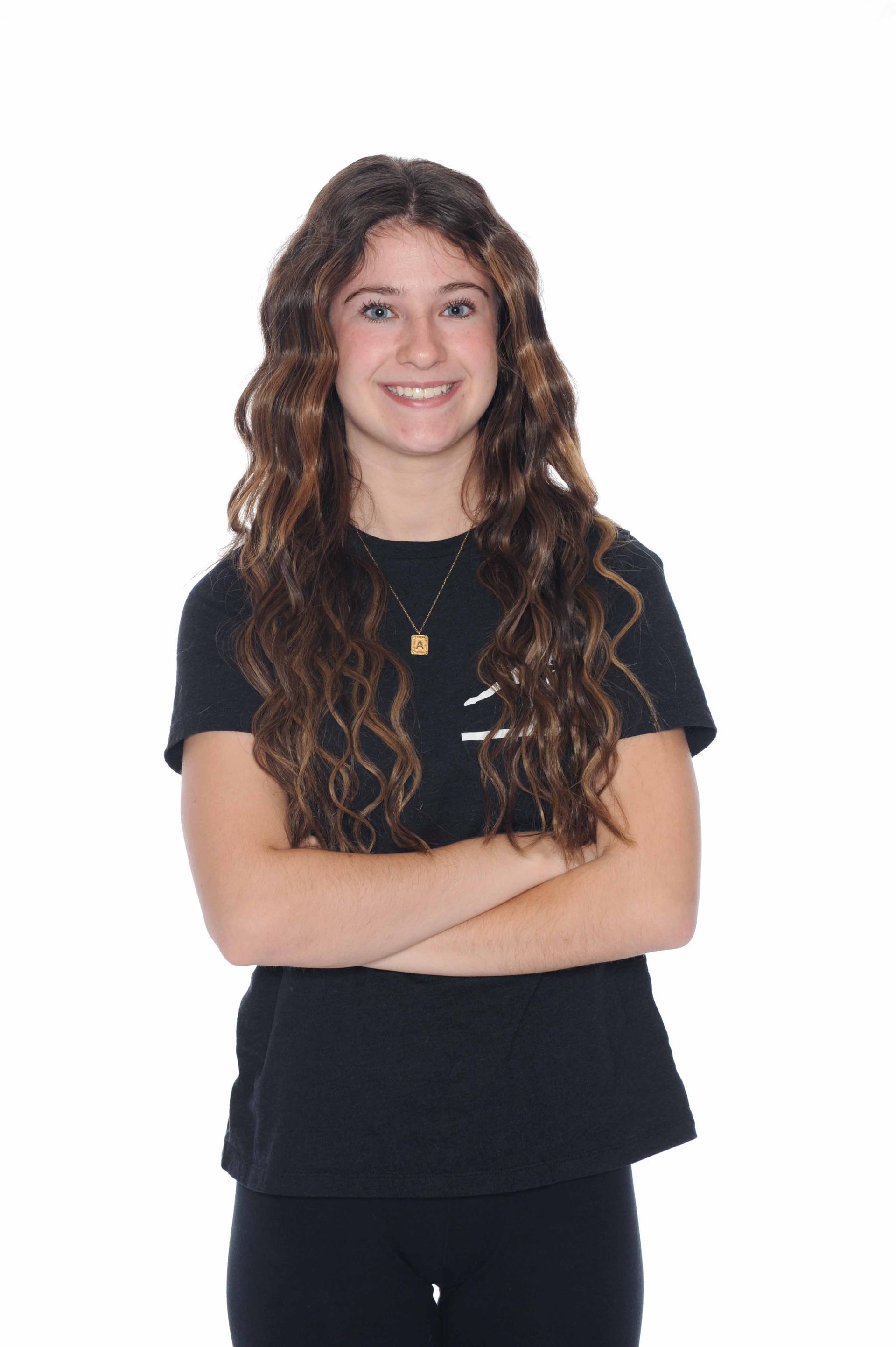 A young woman with long curly hair is standing with her arms crossed and smiling.