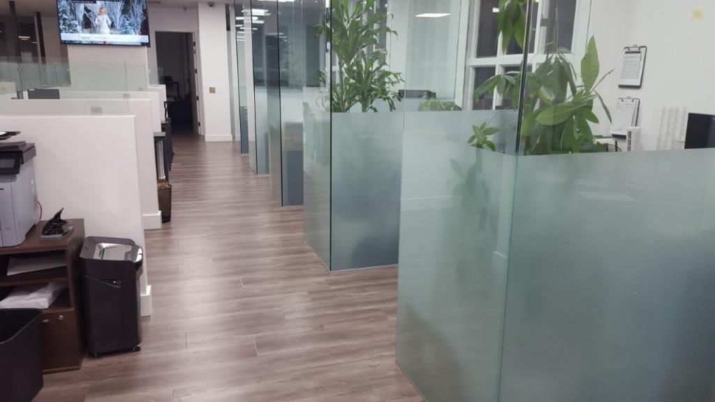 Office hallway with glass dividers, plants, and cubicles. Wooden floor.