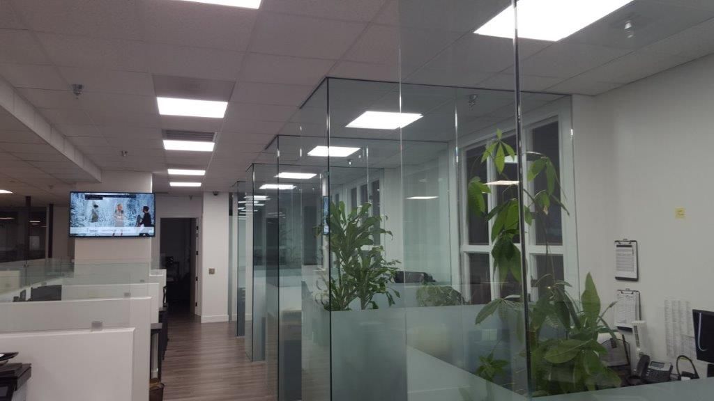 Office interior with glass-walled cubicles and green plants. Fluorescent lights illuminate the space.