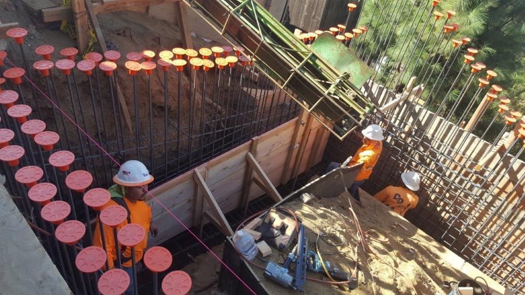 Construction workers pouring concrete in a trench, surrounded by rebar and forms. Orange safety vests and hard hats are visible.