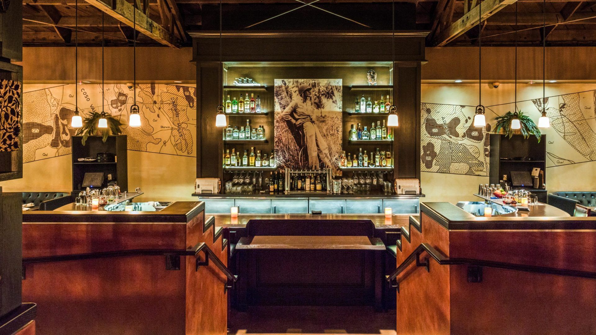 Dimly lit bar with dark wood, stocked shelves, and vintage-style artwork.