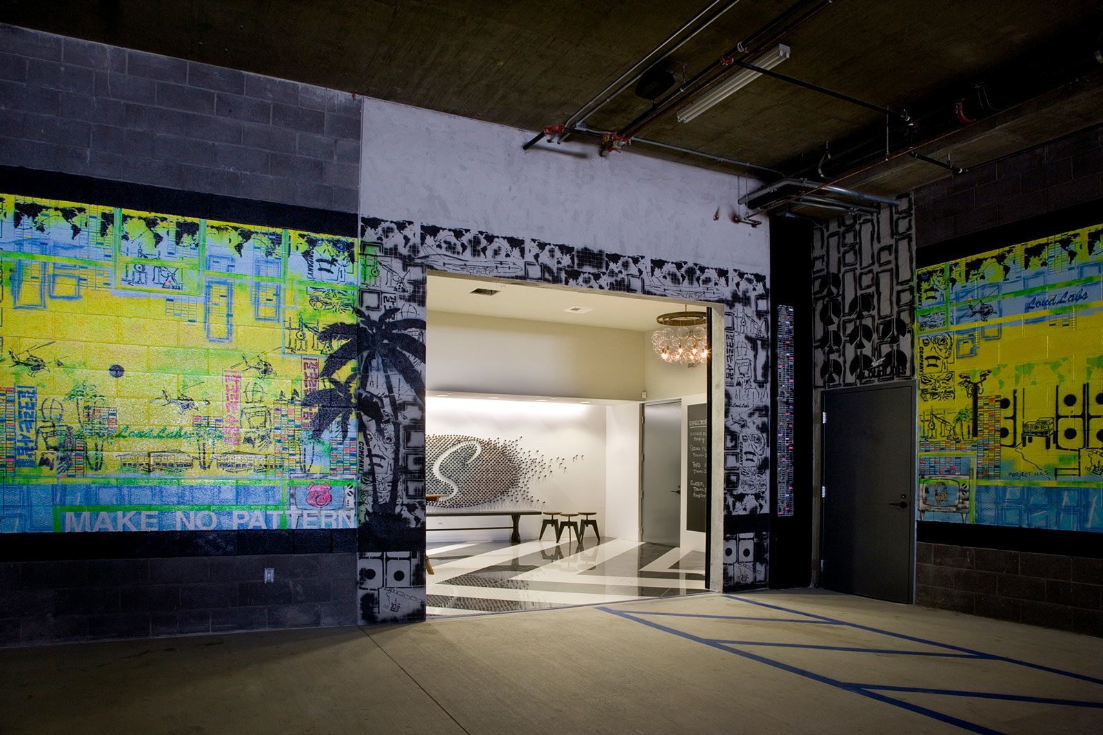 An open doorway leads to a brightly lit space with art. Dark walls with mosaic-like designs.