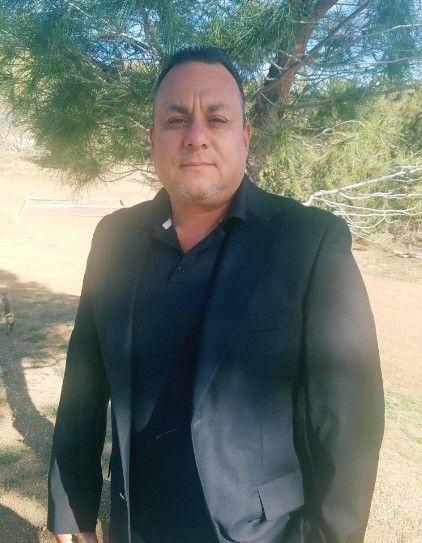 Man in black suit and shirt outdoors, standing in front of a tree, looking at the camera.
