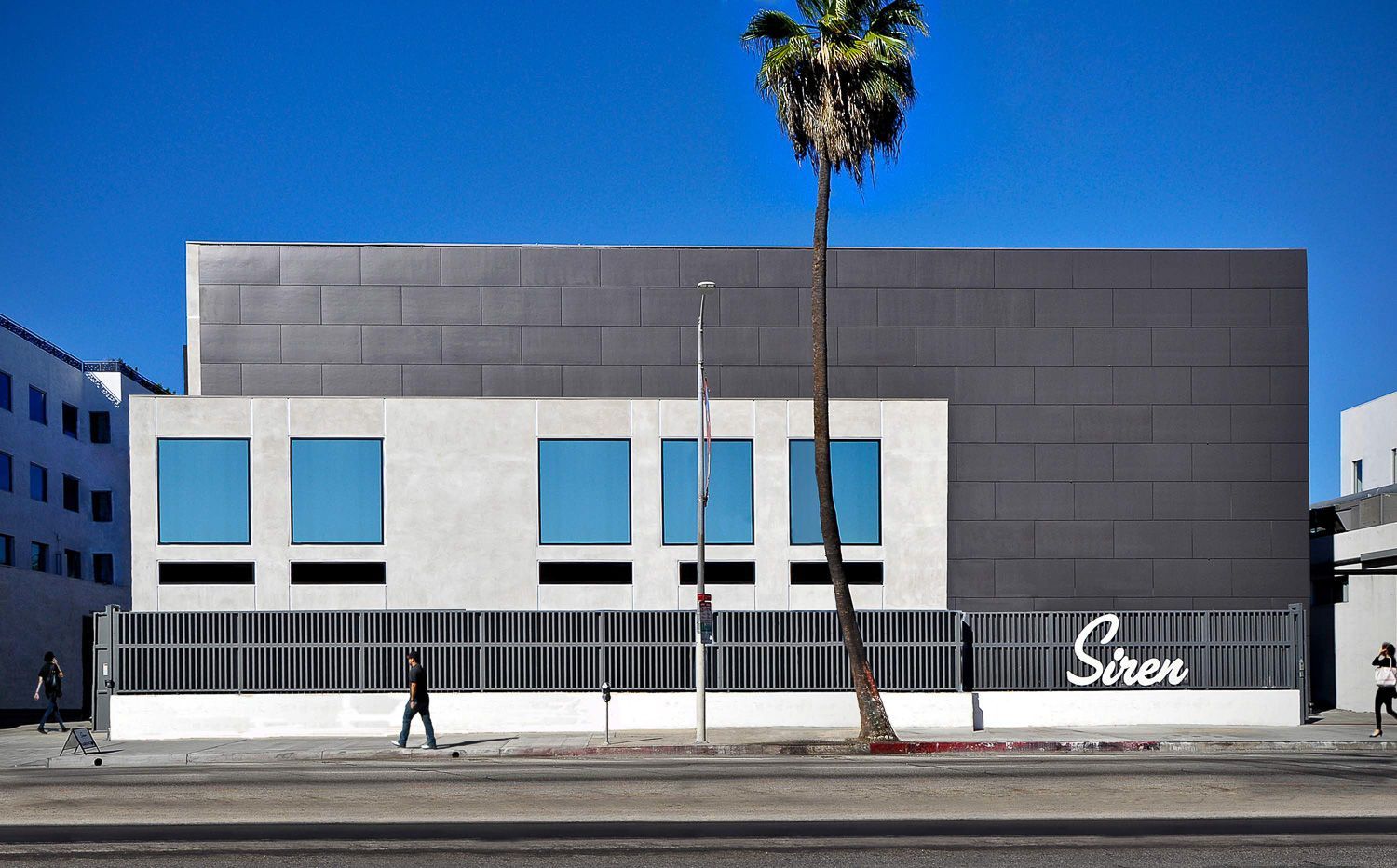 A modern building with a light gray facade, blue windows, and a palm tree in front. A person walks along the sidewalk.