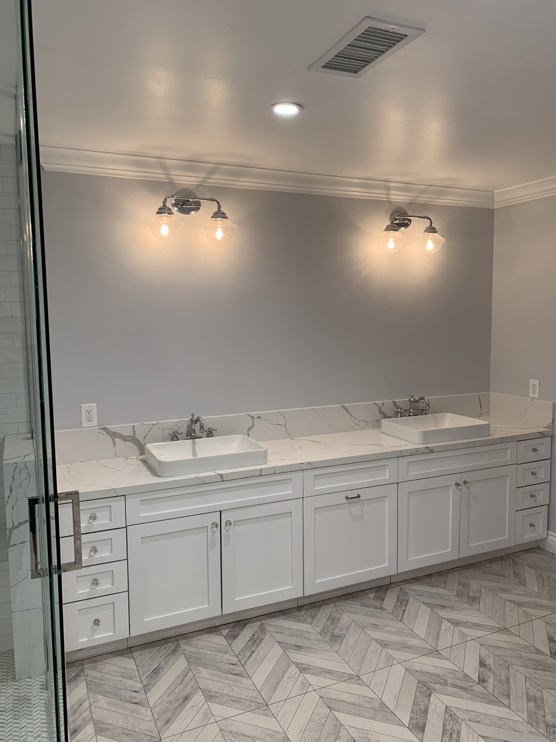Bathroom with double sinks, white cabinets, gray walls, light fixtures, and patterned floor.