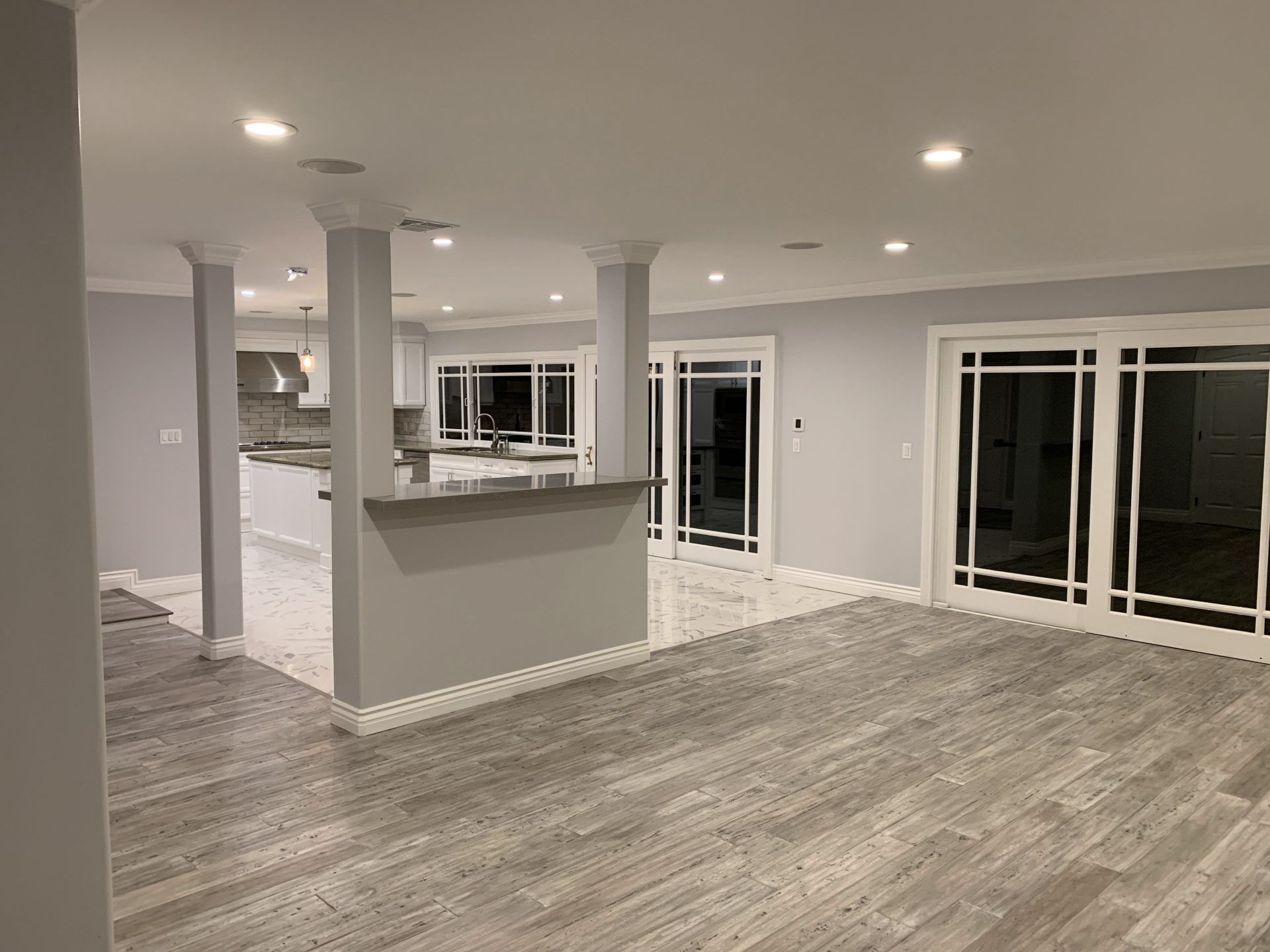 Spacious, newly renovated interior with gray walls, columns, and light wood-look flooring. Kitchen visible in the background.