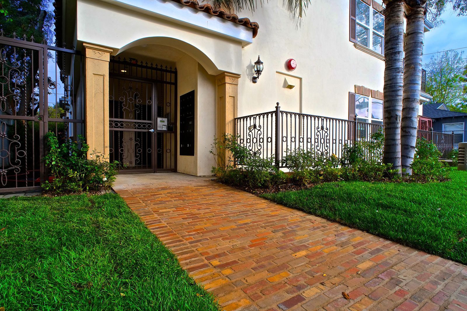 Brick pathway leading to the entryway of a cream-colored building with a wrought-iron gate.