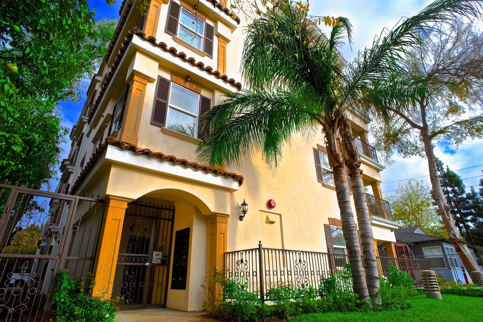 Three-story stucco building with arched entrance, palm trees, and a gated yard.