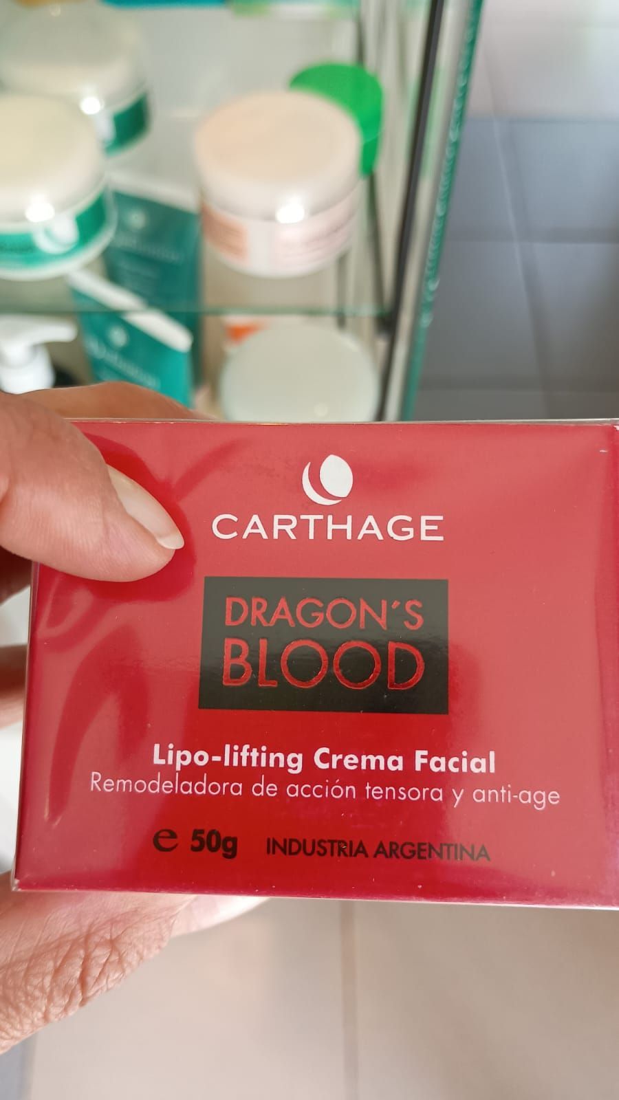 Una mano sostiene una caja roja de crema facial lipolifting Carthago Dragon's Blood de 50 g en un expositor de tienda.