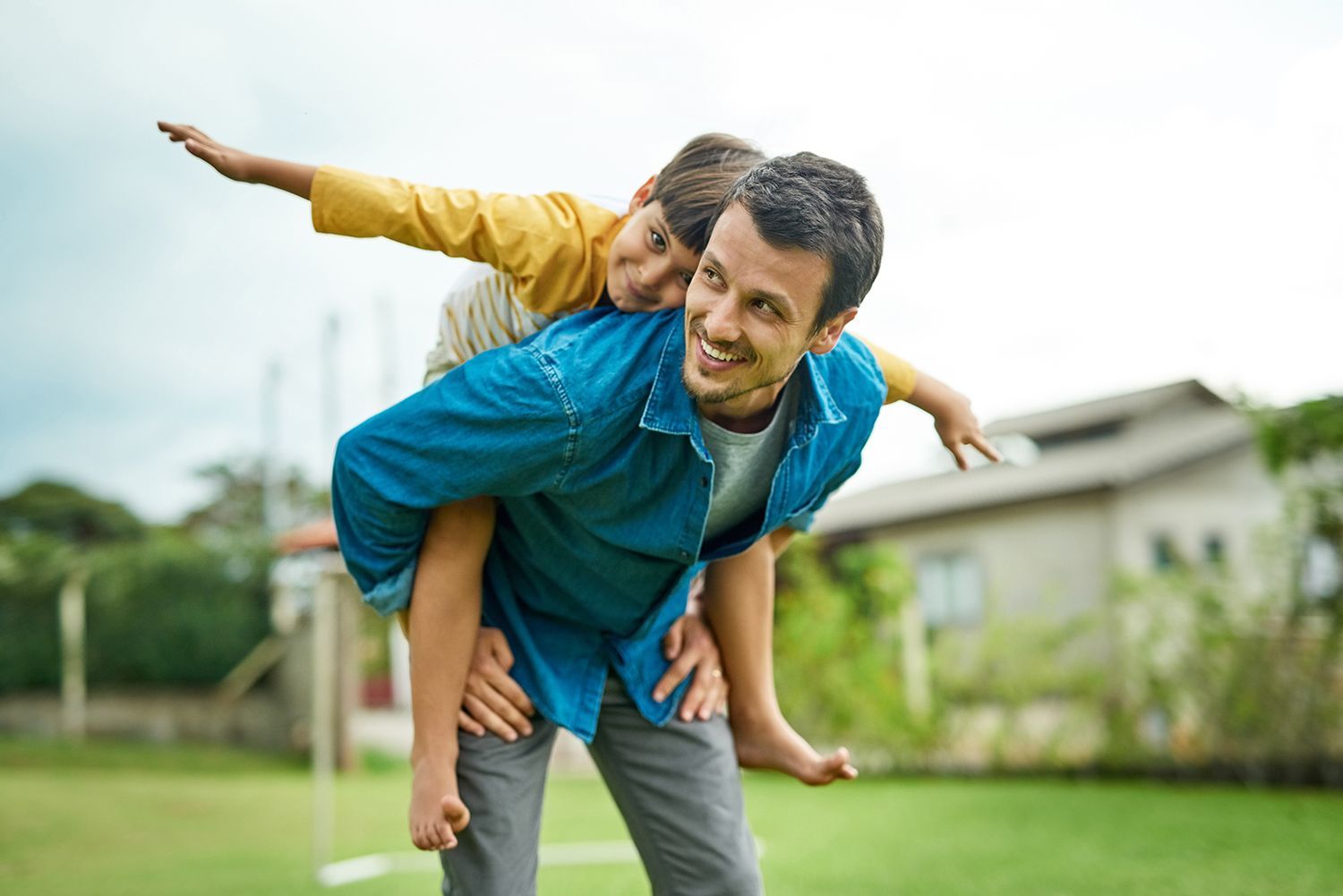 Adult carrying a child on shoulders in a backyard, both posing with outstretched arms.