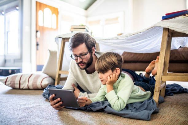 Father and child looking at a tablet under a blanket fort.