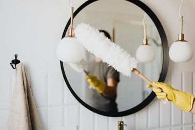 Person dusting a round mirror with a duster, wearing yellow gloves, in a white bathroom.