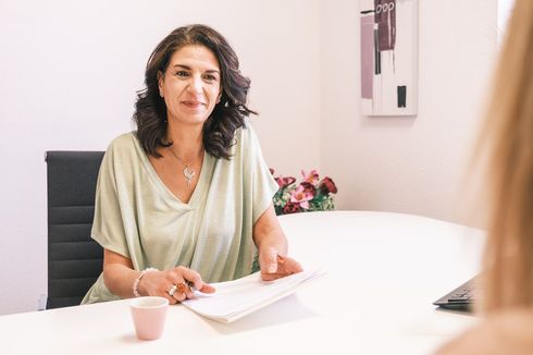 Une femme assise à une table blanche dans une salle de réunion, tenant des papiers et parlant en face de quelqu'un.